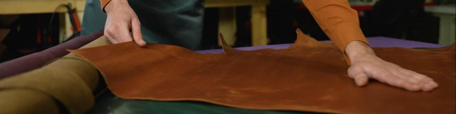 A person's hands smoothing out a large, flat brown leather sheet on a workbench in a workshop setting.